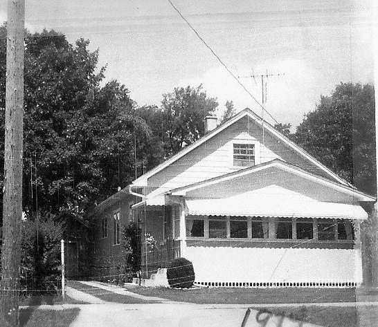 Black-and-white photo of a single-story bungalow with an enclosed front porch, gabled roof, TV antenna, and trees.