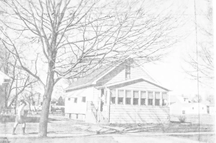 Faded black-and-white photo of a small single-family house with an enclosed front porch, a large leafless tree in the yard, and a person walking on the sidewalk at left.