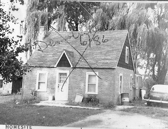 Black-and-white photo of a small brick cottage with steep gable; large X and handwritten "Dec'd 6/86" on roof, car at right, printed caption "HOMESITE" at bottom.