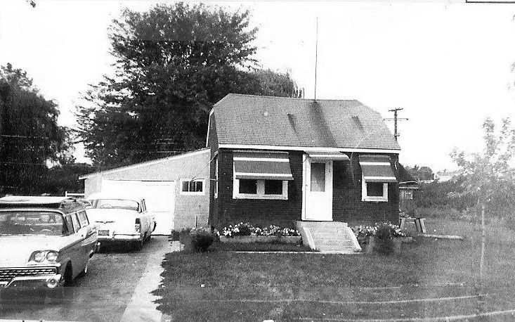 Black-and-white photo of a small gable-roof house with a garage, front lawn, and two vintage cars parked in the driveway.