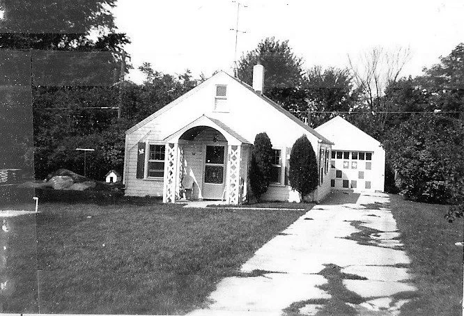 Black-and-white photo of a single-story bungalow with a small covered front porch, lawn, driveway, and detached garage.
