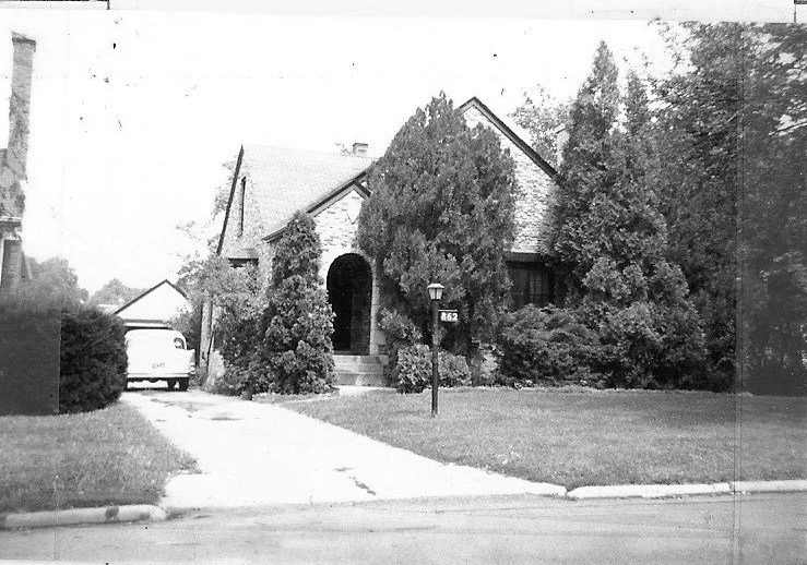 Black-and-white photo of a brick house with an arched front entry surrounded by tall shrubs, a driveway with a parked van, and a mailbox labeled "863."