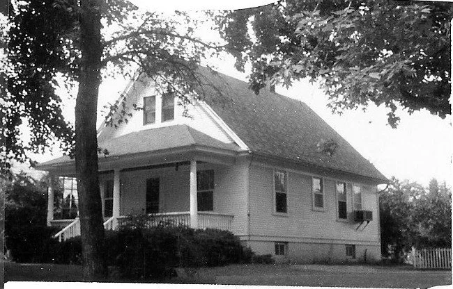 Black-and-white photo of a single-story gabled house with a covered front porch, surrounded by trees and shrubs.