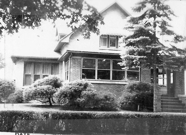 Black-and-white photo of a two-story brick-and-wood house with an enclosed front sunroom, shrubs and evergreen trees in the yard, and a short flight of steps to the right.
