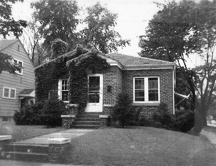 Black-and-white photo of a small brick bungalow with ivy-covered front porch, steps leading to a white door, front lawn, and surrounding trees.