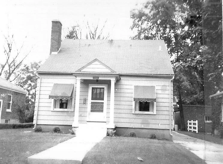 Small single-story house front with central porch and door, two windows with awnings, chimney left and driveway at right.