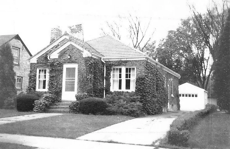 Black-and-white photo of a single-story brick house covered in ivy with a front porch, driveway leading to a detached garage, and a lawn.
