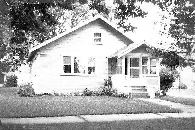 Black-and-white photo of a single-story bungalow with an enclosed front porch and steps, a well-kept lawn and trees, with the house number "120" above the porch.