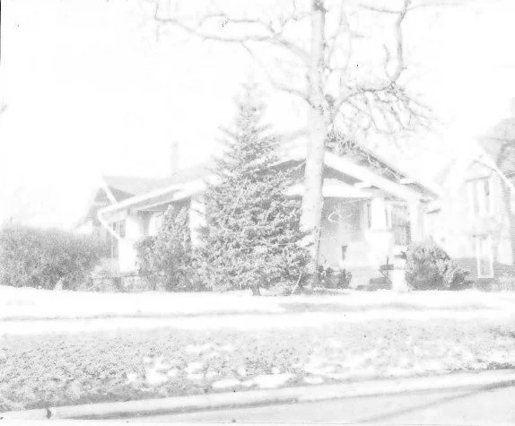 Black-and-white, overexposed photo of a single-story bungalow with a large evergreen and a leafless tree in snow-covered front yard.