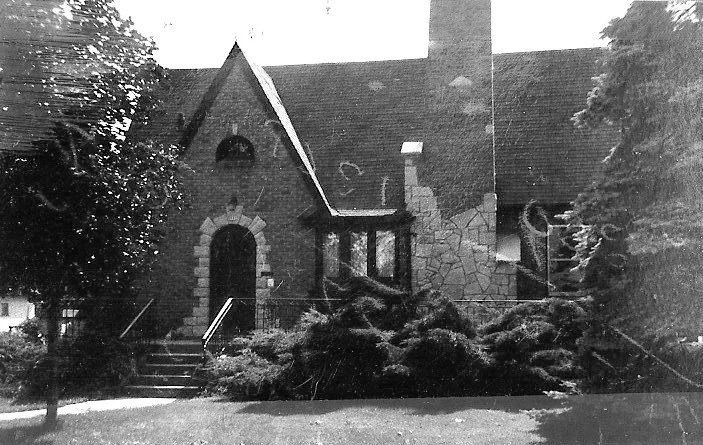 Black-and-white photo of a stone/brick Tudor-style house with steep gable, arched doorway, bay window and dense shrubs in front.