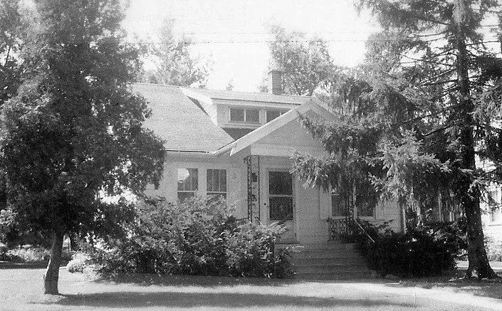 Black-and-white photo of a single-story bungalow with a small front porch, steps, and large trees and shrubs around it.