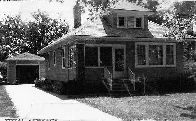 Black-and-white photo of a single-story bungalow with an enclosed front porch, steps up to the entrance, driveway leading to a detached garage, and a front lawn.