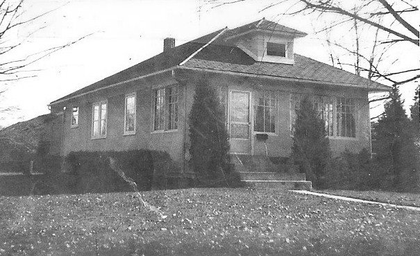 Black-and-white photo of a single-story bungalow with a front porch, steps, shrubs, and a leaf-covered lawn.