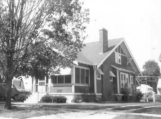 Black-and-white photo of a Craftsman-style bungalow with a covered front porch, chimney, large front-tree, and sidewalk.