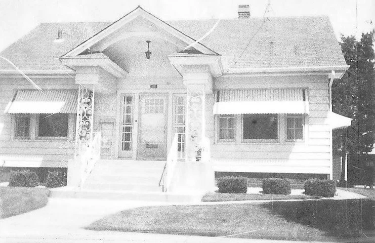 Black-and-white photo of a one-story house with a small covered front porch, decorative columns, concrete steps, window awnings, and trimmed shrubs.
