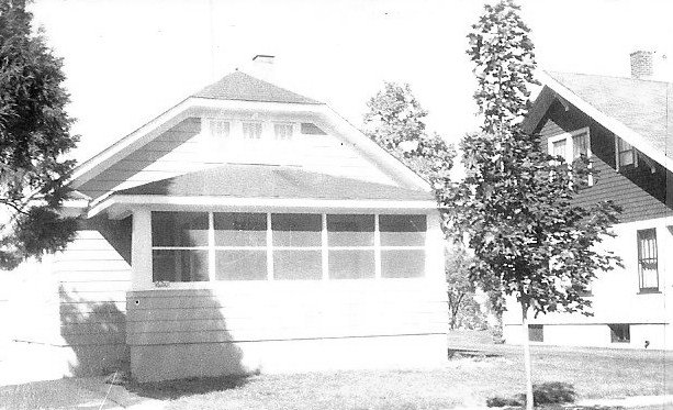 Black-and-white photo of a bungalow-style house with an enclosed front porch and a young tree on the lawn, neighboring house visible.