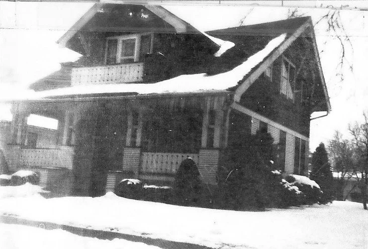 Black-and-white photo of a two-story Craftsman-style house with a covered front porch, dormer windows, and snow on the roof and ground.