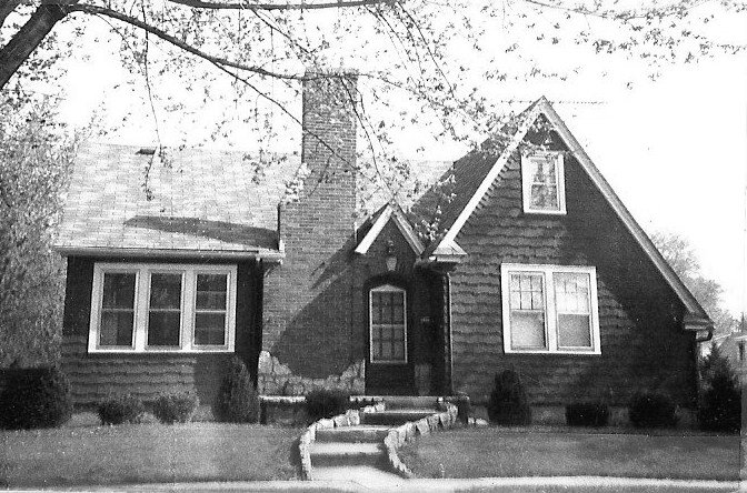 Front view of a small gabled house with a central chimney, stone steps and pathway, and several multi-pane windows.