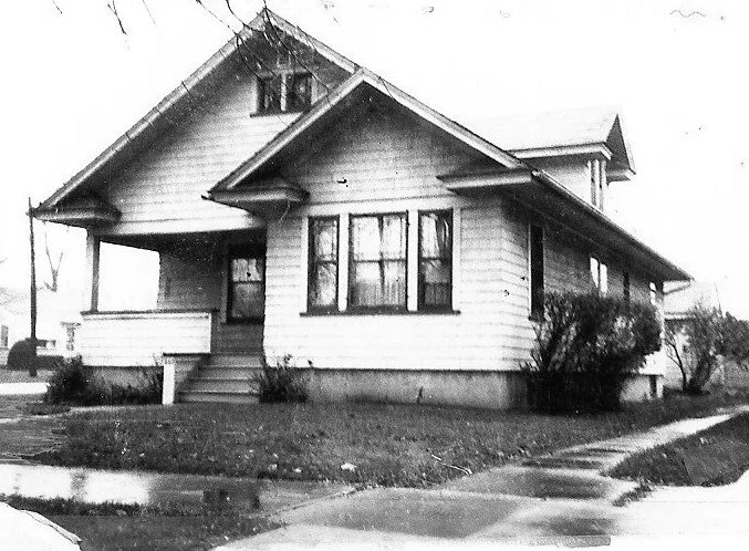 Black-and-white photo of a single-story bungalow with a front porch, steps up to the door, three front windows, and shrubs on a residential corner lot.