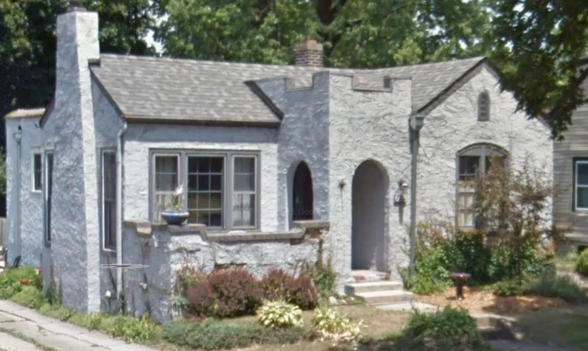 Small light-gray stucco cottage with an arched front doorway, multi-pane window, chimney, and a small landscaped front garden.