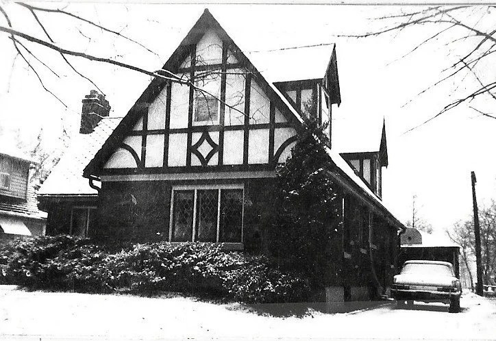 Black-and-white photo of a Tudor-style house with steep gables and half-timbering, snow on the ground and a car parked at the driveway.