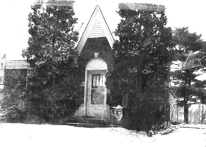 Black-and-white photo of a small brick house with a steep gabled entrance, a centered doorway with decorative fanlight, flanked by large evergreen trees and a snow-covered yard.