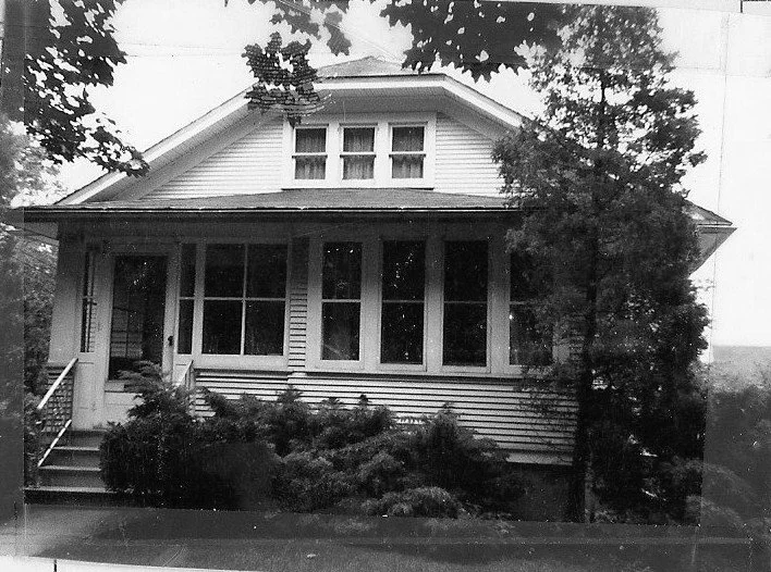 Black-and-white photo of a two-story bungalow with an enclosed front porch, steps, and dense shrubs and trees.