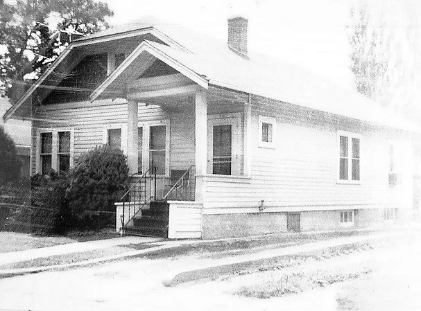 Vintage black-and-white photo of a single-story wood-sided bungalow with a covered front porch, front steps and shrubs.