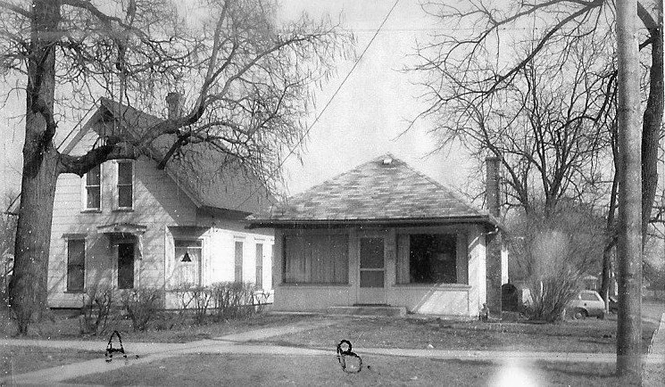 Black-and-white photo of two adjacent houses — a two-story farmhouse and a single-story bungalow — with bare trees and a sidewalk.