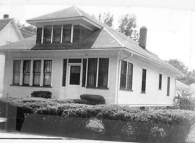 Black-and-white photo of a single-story bungalow with a front porch, dormer windows, chimney, and trimmed hedges.