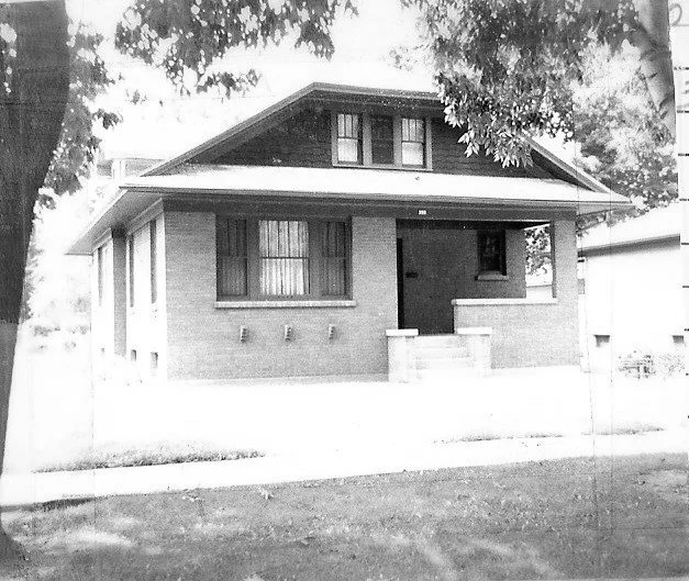Black-and-white photo of a one-story brick bungalow with a covered front porch, small stair, dormer windows, and trees framing the yard.
