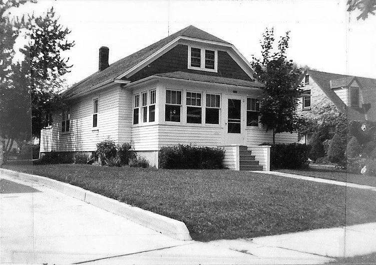 Black-and-white photo of a single-story bungalow with an enclosed front porch, front steps, neatly trimmed lawn and a sidewalk at the corner.
