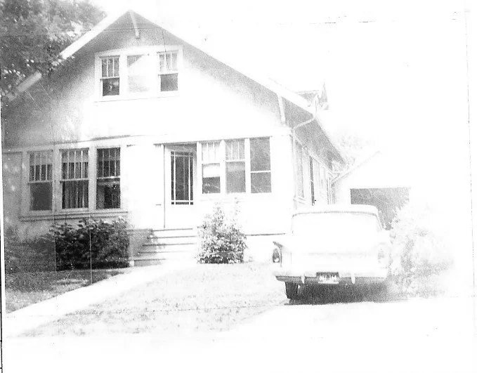 Black-and-white photo of a two-story bungalow with a front porch, shrubs, and a vintage car parked in the driveway.