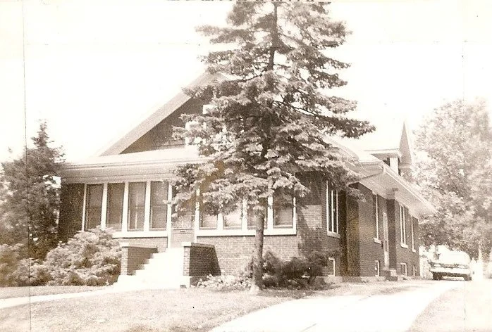 Sepia photo of a brick bungalow with a front porch and steps, a large evergreen tree in front, driveway at right and a parked car.