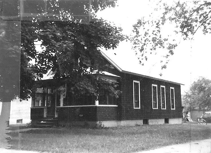 Black-and-white photo of a single-story house with a covered front porch, large tree partly obscuring the façade, and a grassy lawn.