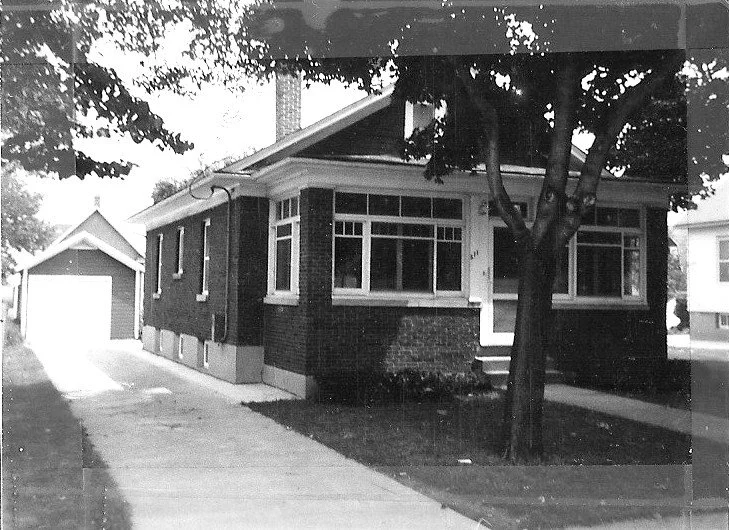 Black-and-white photo of a one-story brick bungalow with an enclosed front porch, tree in the front yard, and a concrete driveway leading to a detached garage.