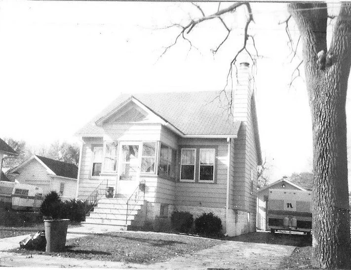 Black-and-white photo of a small single-story house with front steps and enclosed porch, detached garage to the right, and a large tree in the foreground.