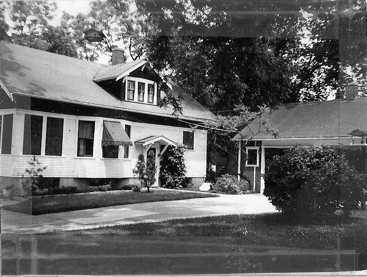 Black-and-white photo of a single-story bungalow with a dormer, small front porch, driveway and attached garage, surrounded by trees and shrubs.