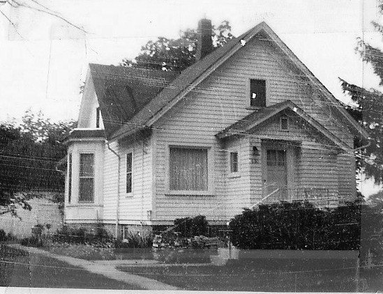 Black-and-white photo of a small wood‑sided gable‑roof house with a front porch, bay window and shrubs.