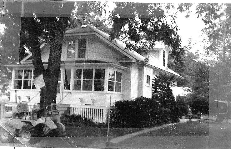 Black-and-white photo of a two-story house with a screened front porch, a large tree in the yard, shrubs along the sidewalk, and an old lawn tractor parked near the curb.
