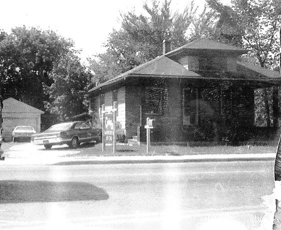 Black-and-white photo of a small brick house with a front porch, mailbox and yard sign, parked cars and trees along a street.
