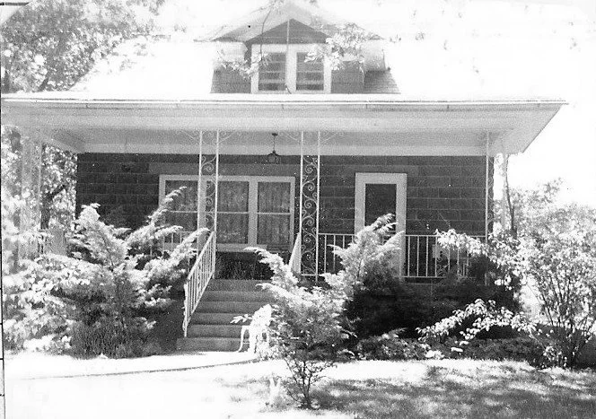 Black-and-white photo of a small front-porch bungalow with steps and decorative metal railings, surrounded by overgrown shrubs and trees.