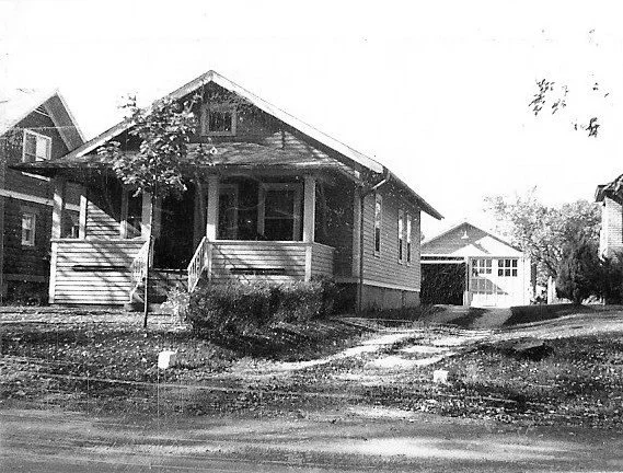 Black-and-white photo of a small single-story bungalow with a covered front porch, yard and a detached garage set back on a slight slope.