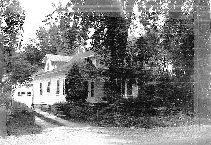 Black-and-white photo of a two-story wooden house with a front porch partly obscured by trees, a driveway leading to a detached garage, and visible scratches/film damage.