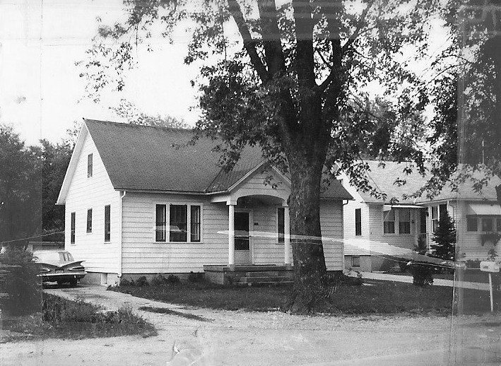 Black-and-white photo of a small single-story clapboard house with a front porch, large tree in the yard and a vintage car parked at the driveway.