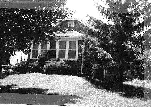 Black-and-white photo of a small bungalow with a screened front porch, surrounded by trees and a grassy front yard.