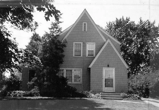 Black-and-white photo of a two-story gabled house with front door, windows, and surrounding trees.