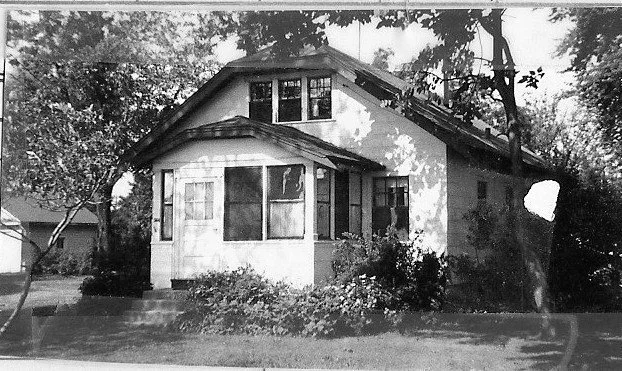 Black-and-white photo of a small two-story bungalow with an enclosed front porch, surrounded by trees and shrubs.