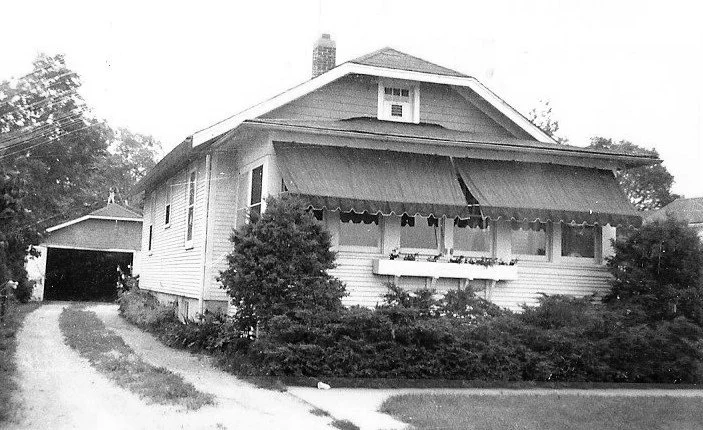 Black-and-white photo of a one-story bungalow with scalloped awnings over the front windows, shrubs, and a driveway leading to a detached garage.