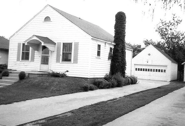 Black-and-white photo of a small single-story white house with a front porch, two shuttered windows, a tall columnar shrub beside it, and a detached garage at the end of a driveway.